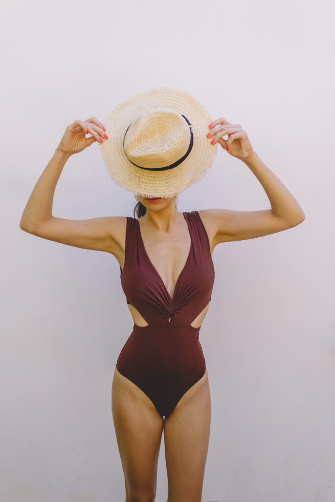 A person in a maroon swimsuit holds a straw hat over their head against a plain background.